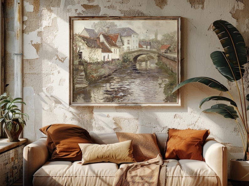Cozy living room with a white sofa adorned with brown and beige cushions. A large painting of a scenic village and bridge above, flanked by lush plants.