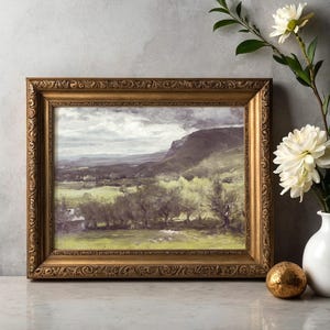 Framed landscape painting of a green valley under a cloudy sky is beside a white vase with white flowers and a small golden sphere on a marble table.