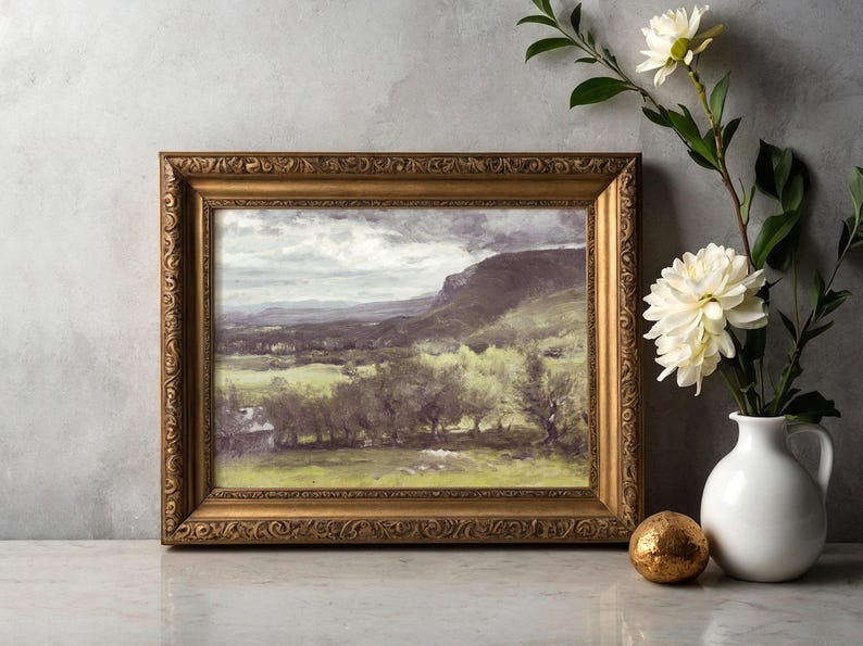 Framed landscape painting of a green valley under a cloudy sky is beside a white vase with white flowers and a small golden sphere on a marble table.