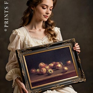 A woman in a vintage white dress holds a framed still-life painting of apples. The setting has a classic, elegant tone with soft lighting.