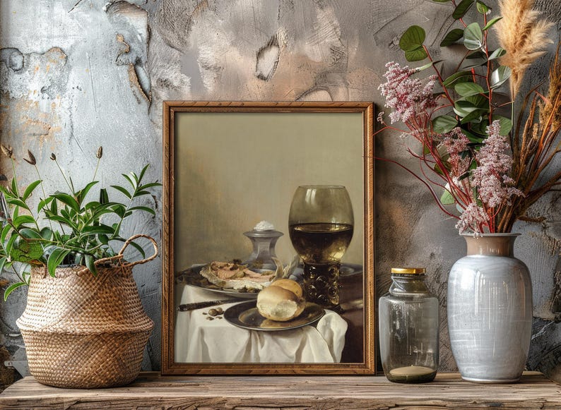 Framed still life of a meal with wine is on a wooden shelf. A woven basket with greenery and a gray vase with pink flowers flank it on either side.