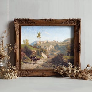 A rustic framed painting depicts a countryside path with donkeys under a blue sky. It rests on a table, flanked by dried flowers in jars, evoking a serene, vintage feel.