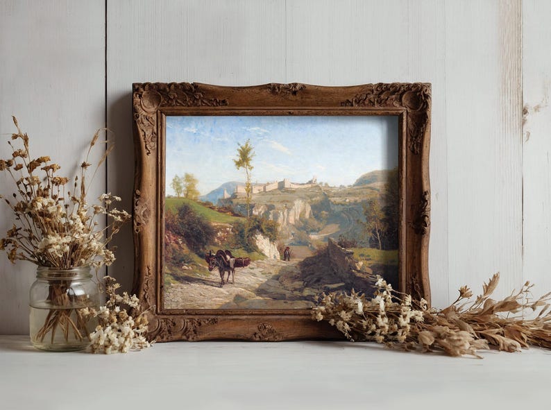 A rustic framed painting depicts a countryside path with donkeys under a blue sky. It rests on a table, flanked by dried flowers in jars, evoking a serene, vintage feel.