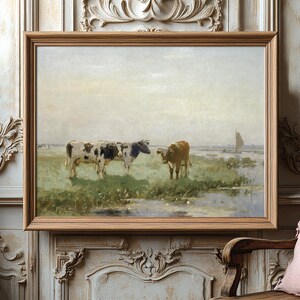 Framed painting of two cows grazing in a serene, grassy field with a misty sky. The artwork is hung on an ornate wall beside a cactus and a cushioned chair.