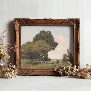 A framed landscape painting of a large, leafy tree with a person walking underneath, set against a partly cloudy sky, displayed on a white wooden surface. To the left of the frame, there is a glass jar filled with dried flowers, and additional dried flowers are scattered on the surface in front of the frame. The background is a white wooden wall.