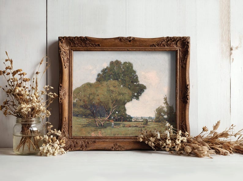 A framed landscape painting of a large, leafy tree with a person walking underneath, set against a partly cloudy sky, displayed on a white wooden surface. To the left of the frame, there is a glass jar filled with dried flowers, and additional dried flowers are scattered on the surface in front of the frame. The background is a white wooden wall.