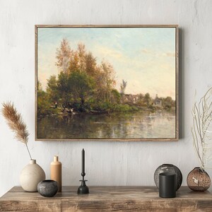 Framed landscape painting of a tranquil river scene on a white wall above a wooden table with minimalist vases and a black candle holder.