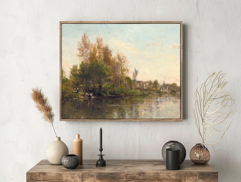 Framed landscape painting of a tranquil river scene on a white wall above a wooden table with minimalist vases and a black candle holder.