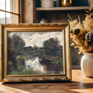 A framed landscape painting of a serene river scene sits on a wooden table beside a vase of dried flowers. Sunlight streams through a window, creating a cozy atmosphere.