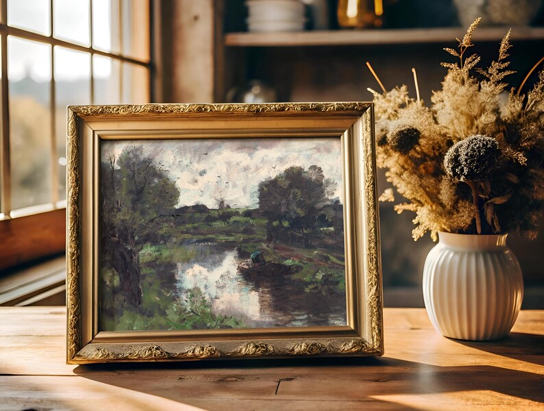 A framed landscape painting of a serene river scene sits on a wooden table beside a vase of dried flowers. Sunlight streams through a window, creating a cozy atmosphere.