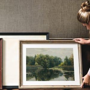 A framed landscape painting with a gold-colored frame, depicting a serene lake scene with trees and reflections. The painting is being held up, with other framed art pieces in the background.