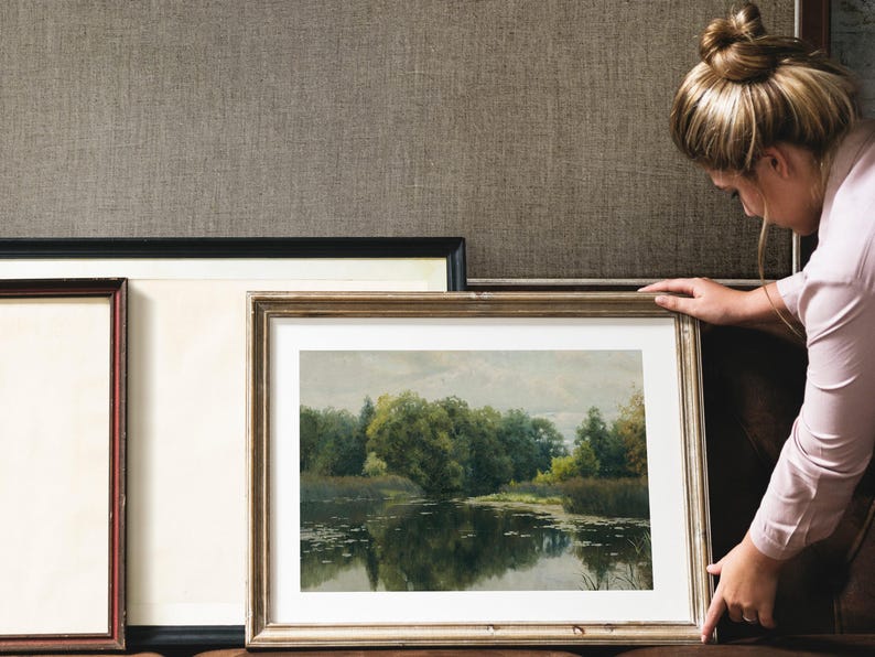 A framed landscape painting with a gold-colored frame, depicting a serene lake scene with trees and reflections. The painting is being held up, with other framed art pieces in the background.