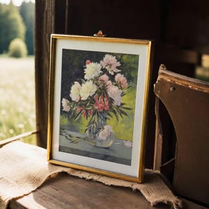 A framed painting of pink and white flowers in a vase sits on wooden surface, with soft outdoor light and blurred green landscape in the background.