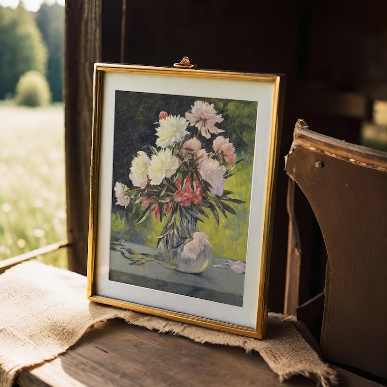 A framed painting of pink and white flowers in a vase sits on wooden surface, with soft outdoor light and blurred green landscape in the background.