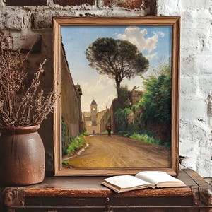 Framed painting on a rustic table against a brick wall, depicting a tree-lined village road. A vase with dried flowers and an open book add charm.