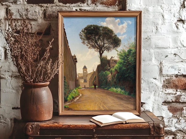 Framed painting on a rustic table against a brick wall, depicting a tree-lined village road. A vase with dried flowers and an open book add charm.