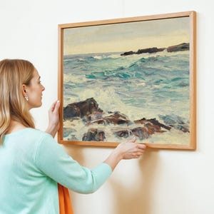 A person in a light blue sweater hangs a framed seascape painting on a white wall. The painting features a turbulent ocean with rocky shores, conveying a serene yet dynamic atmosphere.