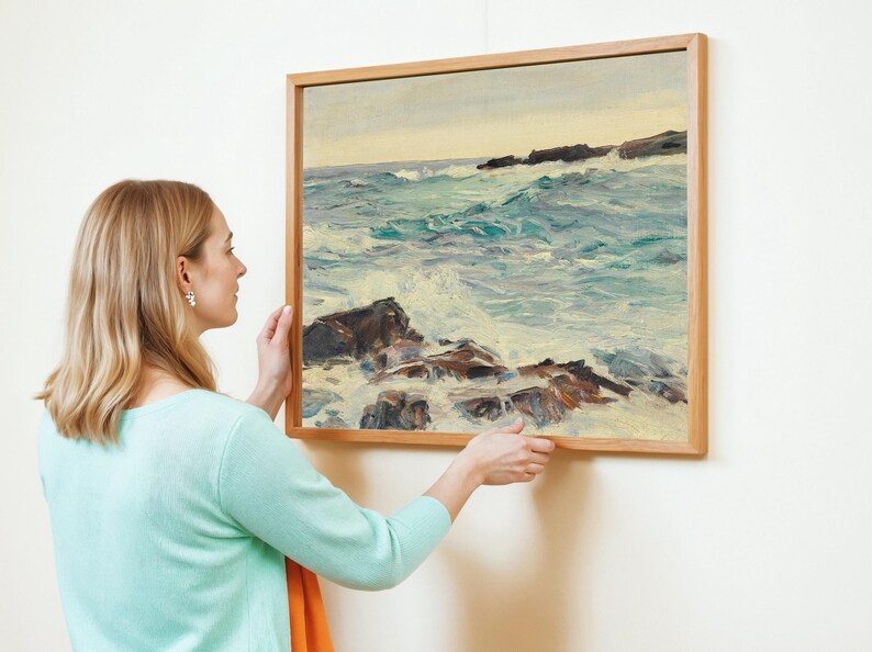 A person in a light blue sweater hangs a framed seascape painting on a white wall. The painting features a turbulent ocean with rocky shores, conveying a serene yet dynamic atmosphere.