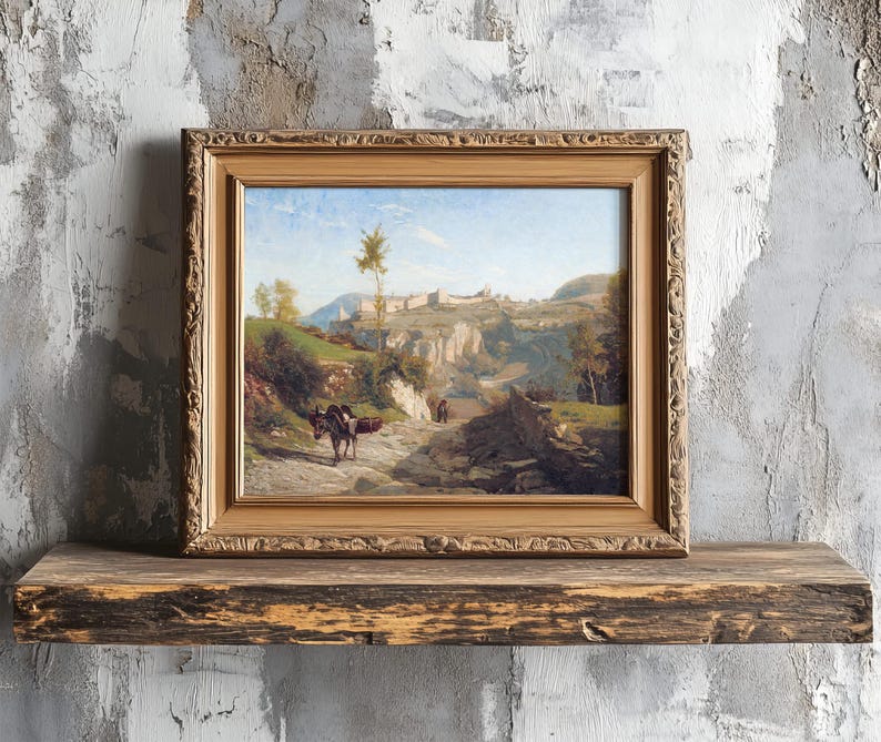 A framed landscape painting on a rustic wooden shelf against a textured wall. It depicts a scenic path with a donkey, leading to distant hills.
