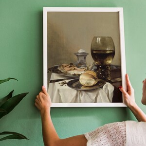 A woman hangs a framed still life painting on a green wall. The artwork features a table with bread, meat, a knife, and a large ornate goblet. A potted plant is visible on the left.