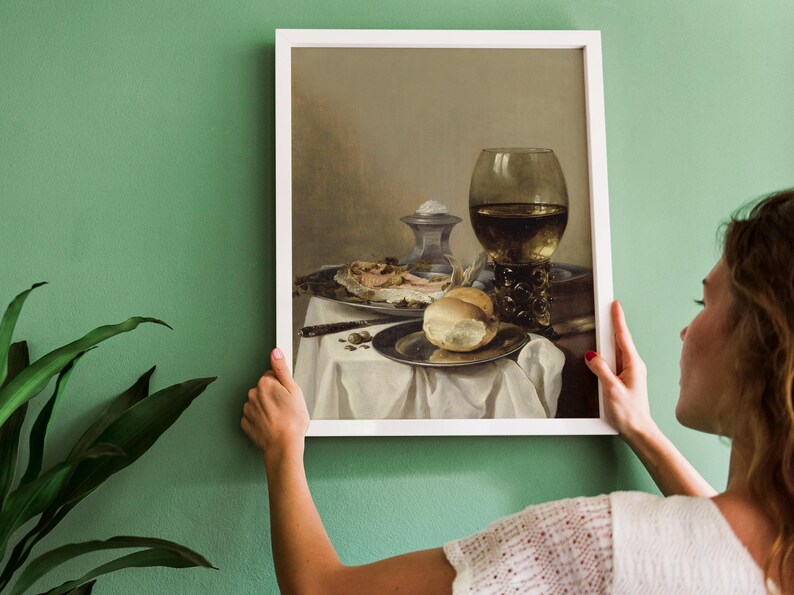 A woman hangs a framed still life painting on a green wall. The artwork features a table with bread, meat, a knife, and a large ornate goblet. A potted plant is visible on the left.
