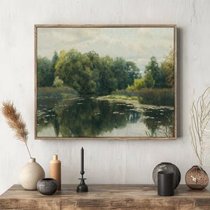 A landscape painting of a tranquil pond surrounded by lush trees is framed on a textured wall above a wooden table adorned with minimalist vases and dried plants.
