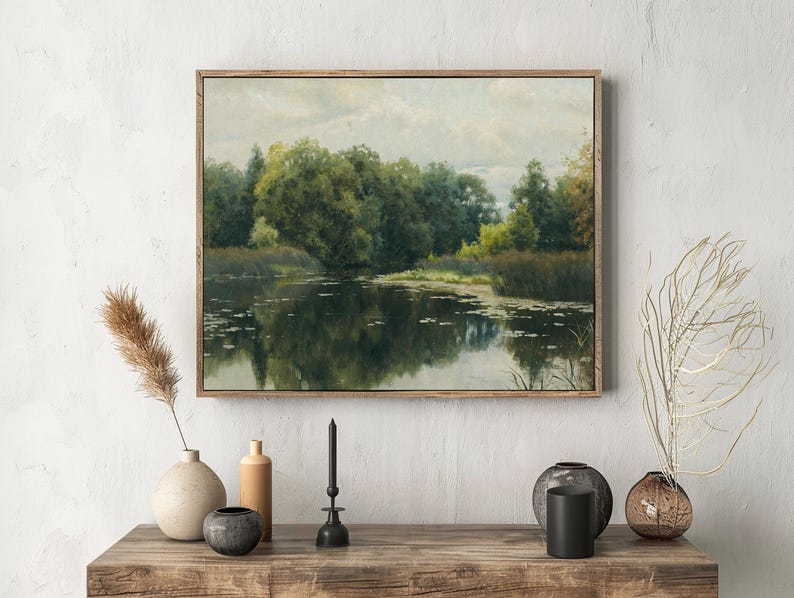 A landscape painting of a tranquil pond surrounded by lush trees is framed on a textured wall above a wooden table adorned with minimalist vases and dried plants.
