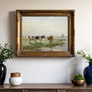 A framed painting of cows in a field hangs on a wall. Below, a wooden console holds a black vase with greenery, a white jar, and a blue vase with white tulips.