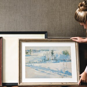 A woman in a pastel shirt carefully adjusts a framed winter landscape painting against a textured wall, conveying a sense of contemplation and care.