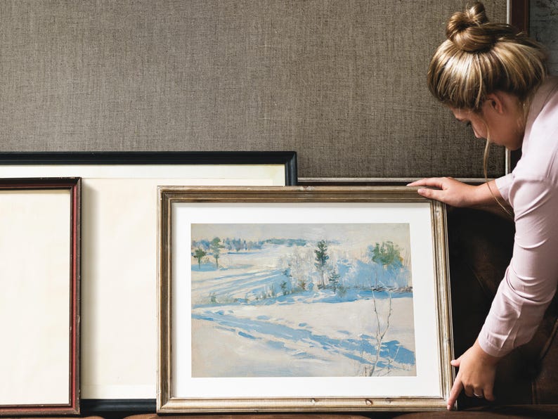 A woman in a pastel shirt carefully adjusts a framed winter landscape painting against a textured wall, conveying a sense of contemplation and care.