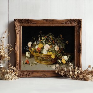 A framed painting of a basket filled with colorful flowers, set against a dark background. Rustic wood frame, surrounded by dried flowers on a white surface.