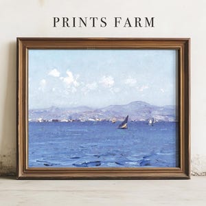 A framed seascape painting leaning against a wall, featuring a sailboat on a calm blue sea with mountains and clouds in the background. The words "PRINTS FARM" are above it.
