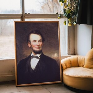 A framed portrait of a bearded man in a suit leans against a window with a scenic view. Nearby, a cozy yellow chair adds warmth to the setting.