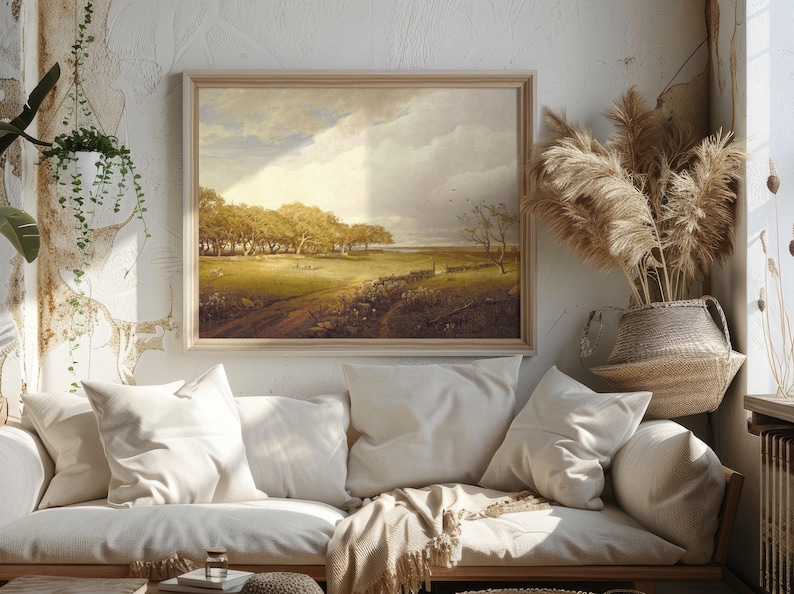 Cozy living room with a white sofa adorned with soft pillows and blankets beneath a landscape painting. Sunlight filters through, highlighting pampas grass decor.