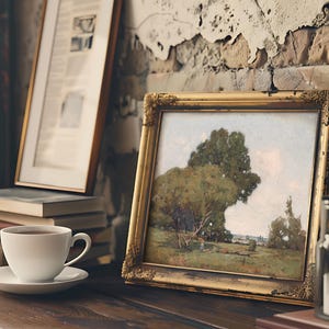 A cozy scene featuring a framed landscape painting of a large tree and open fields, placed on a rustic wooden table against a textured, peeling brick wall. In front of the painting, there is a white cup filled with coffee on a matching saucer, with a stack of books beside it. A vintage lantern is also partially visible on the right side of the image.