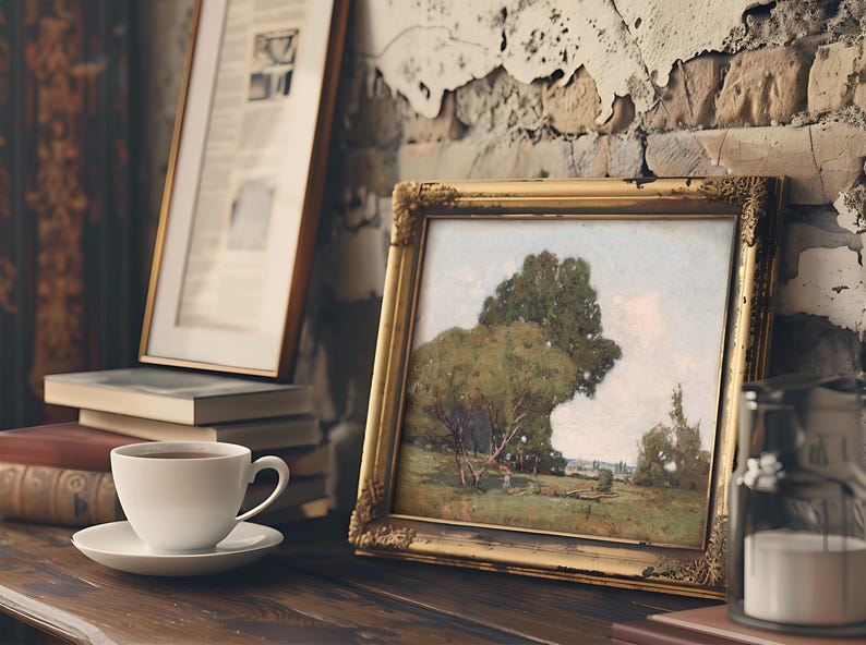 A cozy scene featuring a framed landscape painting of a large tree and open fields, placed on a rustic wooden table against a textured, peeling brick wall. In front of the painting, there is a white cup filled with coffee on a matching saucer, with a stack of books beside it. A vintage lantern is also partially visible on the right side of the image.