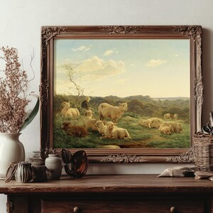 A framed painting on a rustic wooden table shows sheep in a lush field under a blue sky. Nearby, dried flowers and artist tools add a warm, pastoral feel.