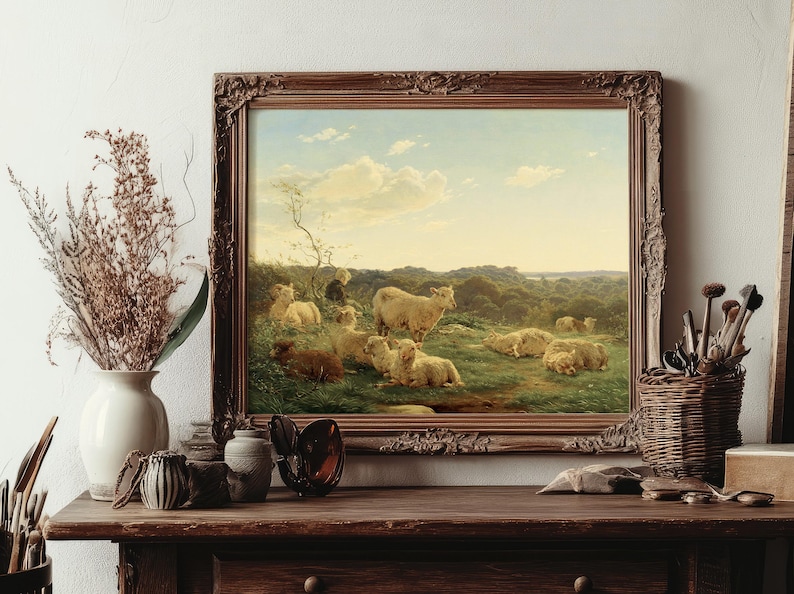 A framed painting on a rustic wooden table shows sheep in a lush field under a blue sky. Nearby, dried flowers and artist tools add a warm, pastoral feel.