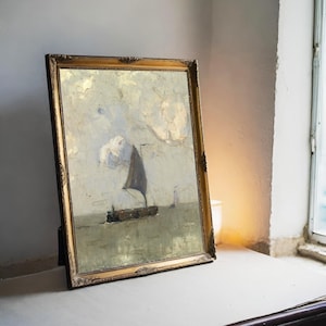 A framed painting of a sailboat on a calm sea leans against a wall on a table. Soft lighting from a nearby window creates a serene atmosphere.