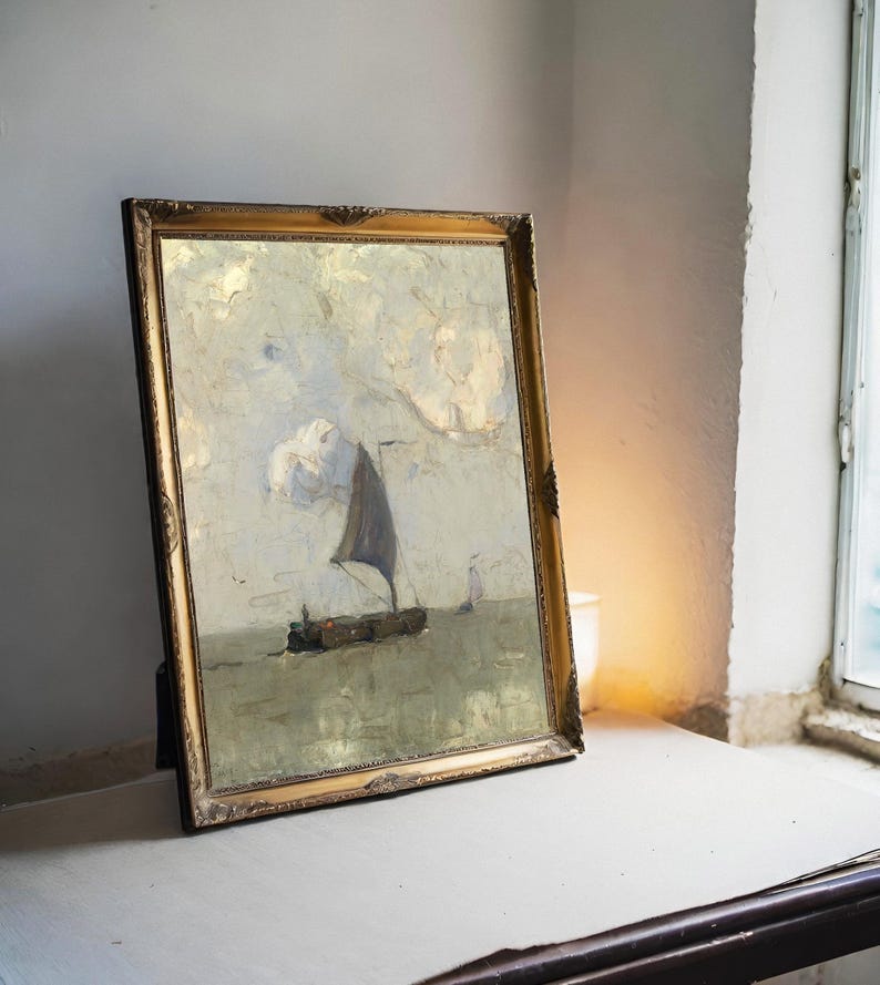 A framed painting of a sailboat on a calm sea leans against a wall on a table. Soft lighting from a nearby window creates a serene atmosphere.