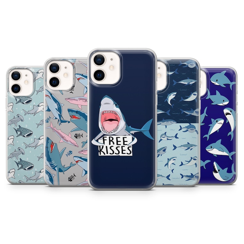 Pu&ograve; includere: Cinque diverse custodie per iPhone con design a squalo. Le custodie sono trasparenti e mostrano il design del telefono. Una custodia presenta uno squalo con il testo "Free Kisses". Un'altra custodia presenta un motivo di squali e pesci. Le custodie rimanenti presentano diversi design di squali.
