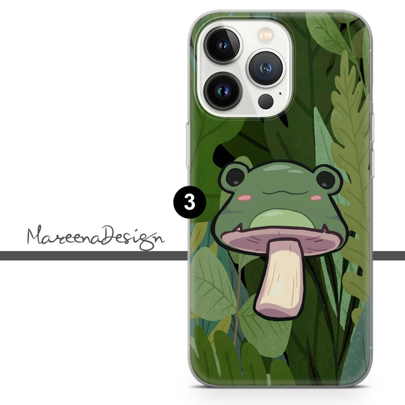 Op de afbeelding: Een transparante telefoonhoes met een groene kikker die op een paddenstoel zit. De kikker heeft een groen lichaam, witte ogen en een roze mond. De achtergrond is een groen en bruin bladmotief.