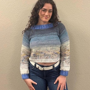 May include: A cropped, hand-crocheted sweater in a gradient of blues, grays, and creams. The sweater features a ribbed neckline and cuffs in a solid blue. A white belt cinches the waist, paired with blue jeans.