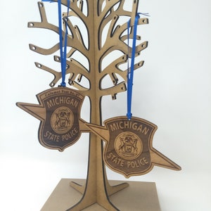 May include: Two wooden ornaments shaped like a police badge with the text "Michigan State Police" and the state seal. The ornaments are hanging from a wooden tree-shaped stand with blue ribbon.
