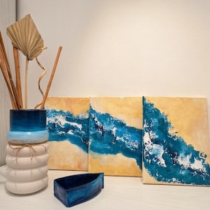 Könnte beinhalten: Eine zylindrische Vase mit geripptem weißem Fuß und blauem Farbverlauf am oberen Rand, gefüllt mit getrockneten Pflanzenstängeln. Daneben stehen eine kleine blaue bootsförmige Schale und drei rechteckige Leinwände mit abstrakten blauen Meereswellen.