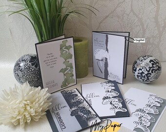 May include: Five greeting cards with a black and white ginkgo leaf design. The cards are on a white surface with a green plant, a white flower, and two decorative bowls.