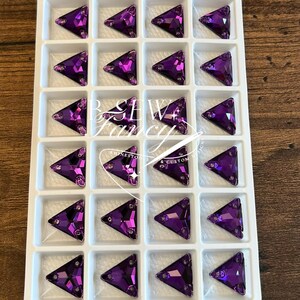Pode incluir: Uma bandeja de strass roxos triangulares e facetados. Cada strass está colocado em um compartimento de plástico branco. Os strass têm uma ponta e são dispostos em um padrão de grade. A imagem é vista de cima.