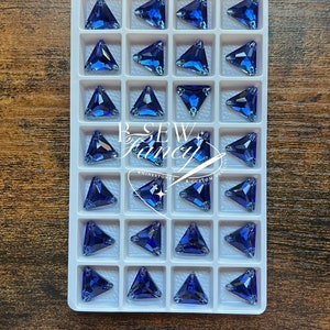 Op de afbeelding: Een dienblad gevuld met driehoekige, donkerblauwe strass-steentjes. Elke edelsteen is gezet in een wit plastic raster. De strass-steentjes zijn gerangschikt in een rasterpatroon. De tray staat op een houten oppervlak.