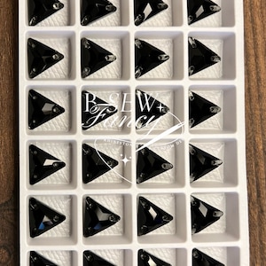 May include: A tray of black triangular rhinestones. Each rhinestone has a faceted surface and is set with small silver prongs. The rhinestones are arranged in a grid pattern within a white plastic tray. The tray is on a wooden surface. The text "B SEW + Fancy" is visible.