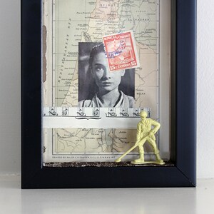 May include: A framed collage with a vintage map of Palestine, a black and white photo of a woman, a piece of sheet music, and a green plastic toy soldier. The map has the text "MODERN PALESTINE" and "REVISED BY MAJOR CR CONDER D.C.L.L.O.M.R.A.S.R.E.". The stamp on the map reads "15 CENTIMOS 15".